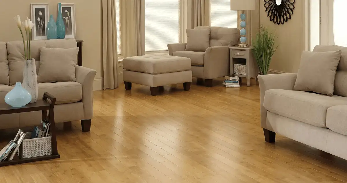 A living room with professionally installed bamboo flooring, beige sofas, a matching ottoman, a side table with a blue lamp, vases, framed pictures, and large windows with blinds and curtains in Lowell, MA