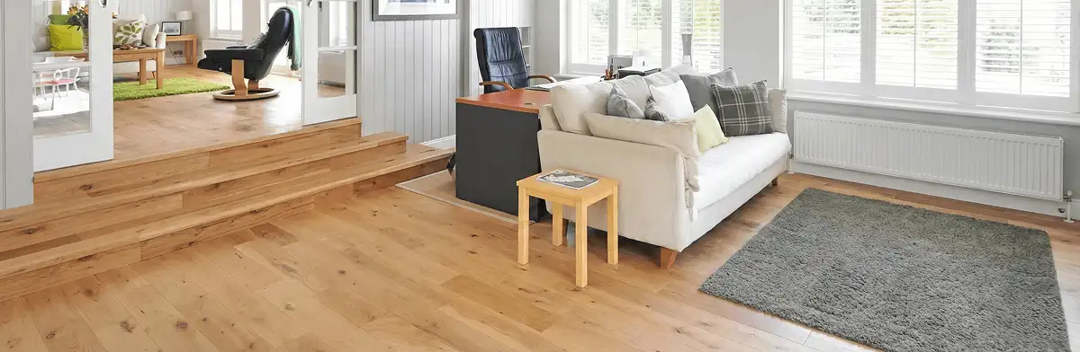 A modern living room with professionally installed laminate flooring, a beige sofa, a small side table, a gray rug, and large windows with white shutters in Lowell, MA